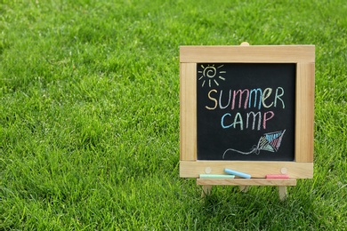 Photo of Blackboard with text SUMMER CAMP and drawings on green grass. Space for text