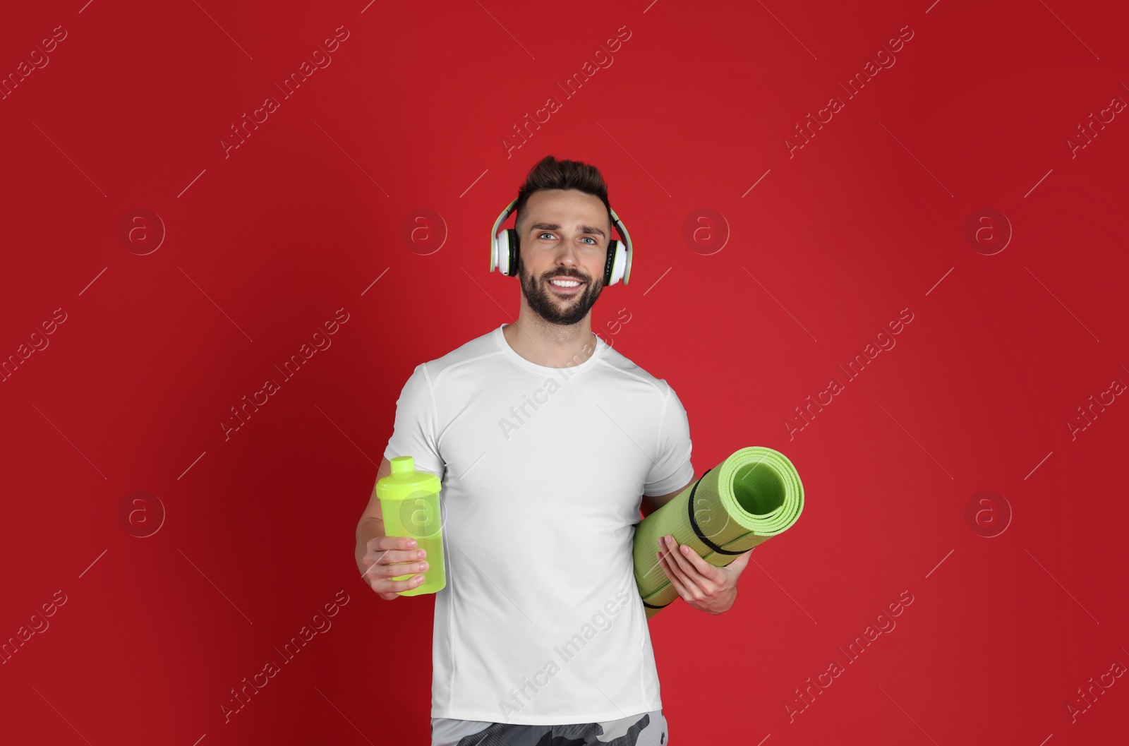 Photo of Handsome man with headphones,yoga mat and shaker on red background