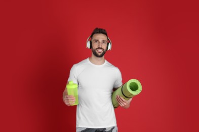 Handsome man with headphones,yoga mat and shaker on red background
