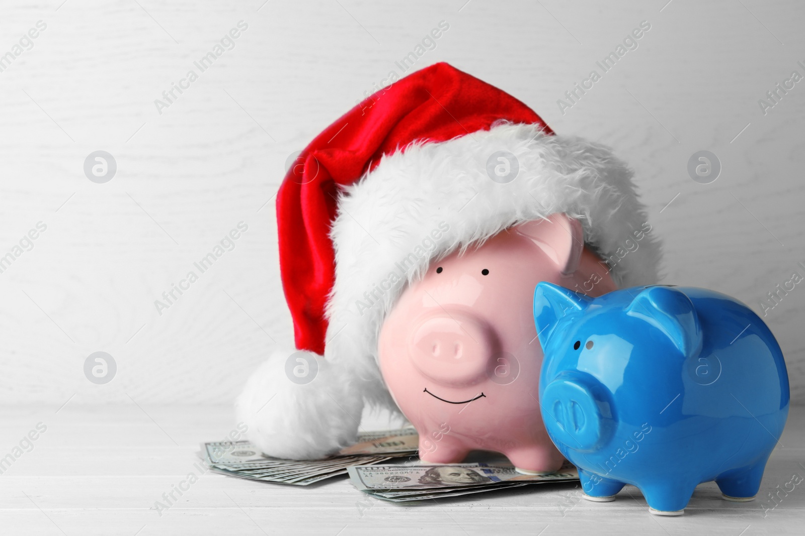 Photo of Piggy banks with Santa hat and dollar banknotes on white wooden table. Space for text