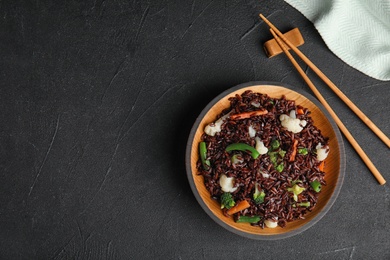 Photo of Plate of brown rice with vegetables and chopsticks on black table, flat lay. Space for text