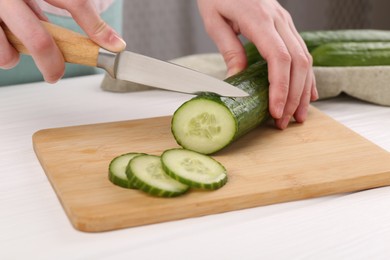 Woman cutting cucumber on wooden board at white table, closeup