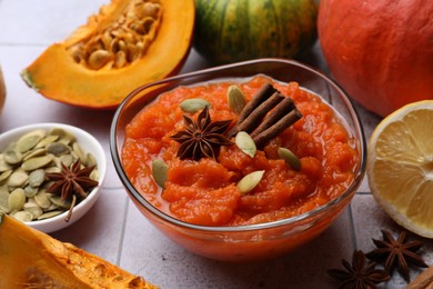 Photo of Bowl of delicious pumpkin jam and ingredients on tiled surface, closeup