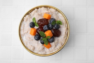 Photo of Delicious barley porridge with blueberries, pumpkin, dates and mint in bowl on white tiled table, top view