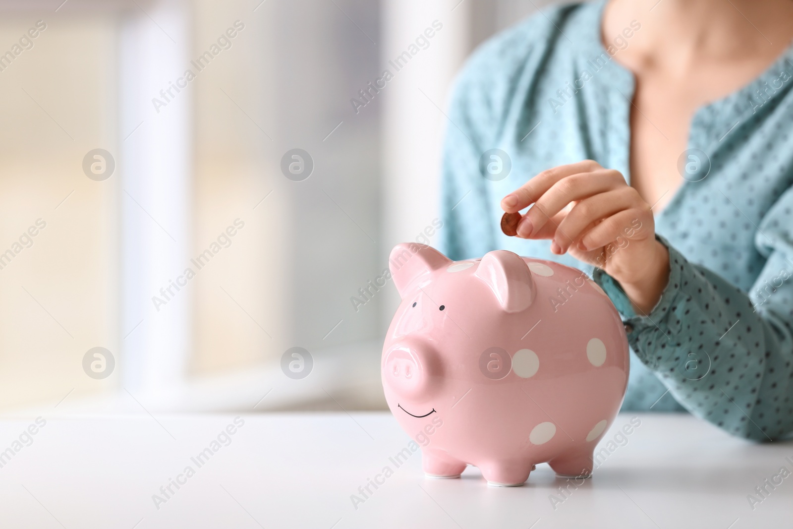 Photo of Woman putting coin into piggy bank at table indoors, closeup. Space for text