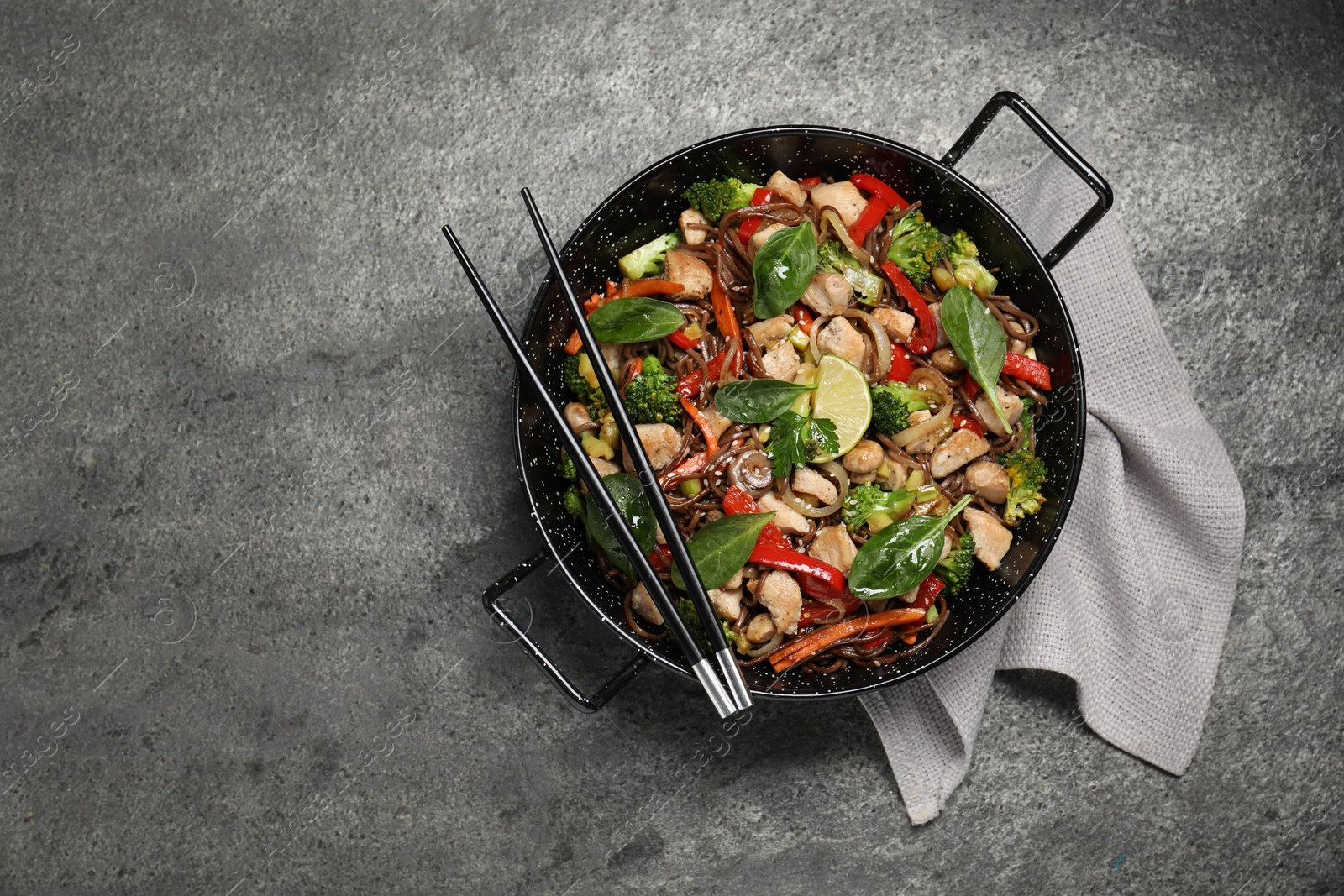 Photo of Stir-fry. Tasty noodles with meat in wok and chopsticks on grey textured table, top view