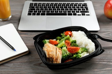 Photo of Container with tasty food, notebook and laptop on wooden table. Business lunch