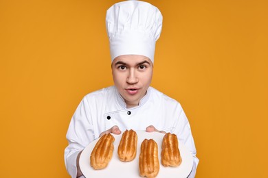 Photo of Portrait of happy confectioner in uniform holding plate with eclairs on orange background