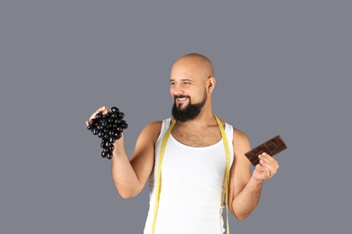 Overweight man with grapes, chocolate bar and measuring tape on gray background