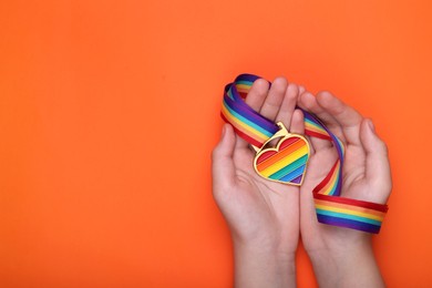 Photo of Woman holding rainbow ribbon with heart shaped pendant on orange background, top view and space for text. LGBT pride