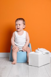 Little child sitting on baby potty and box with diapers near orange wall