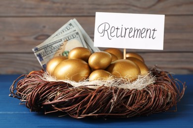 Photo of Golden eggs, dollar banknotes and card with word Retirement in nest on blue wooden table. Pension concept