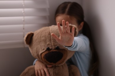 Photo of Child abuse. Little girl with teddy bear doing stop gesture indoors, selective focus
