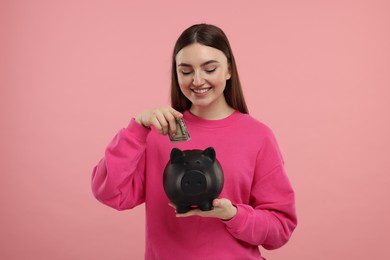 Happy woman putting dollar banknote into piggy bank on pink background