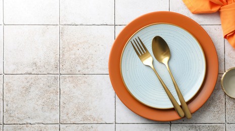 Stylish setting with cutlery, napkin and plates on light tiled table, top view. Space for text