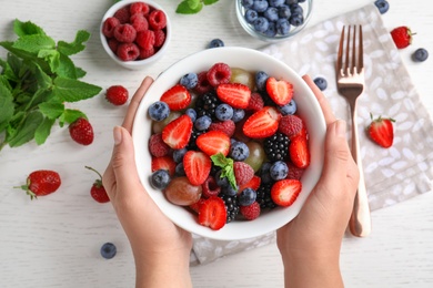 Photo of Woman holding bowl with fresh tasty fruit salad over white wooden table, top view