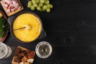 Photo of Fondue pot with melted cheese, fork and different products on black wooden table, flat lay. Space for text