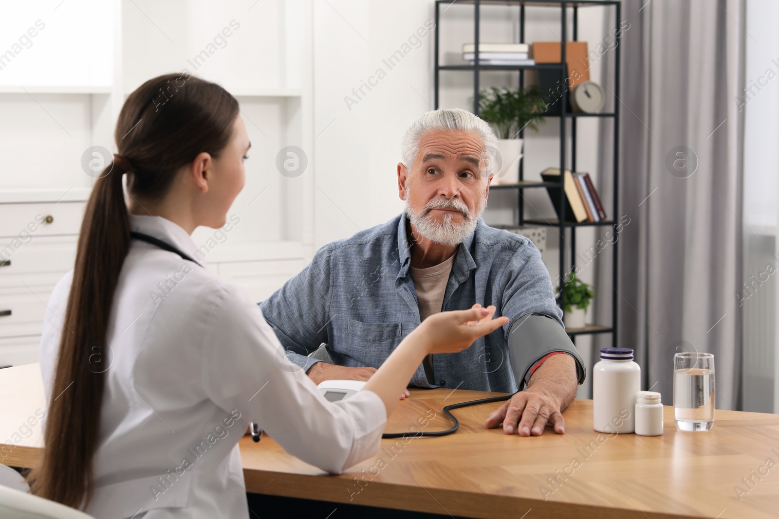 Photo of Young healthcare worker measuring senior man's blood pressure at wooden table indoors