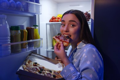 Photo of Young woman eating pizza near refrigerator in kitchen at night. Bad habit