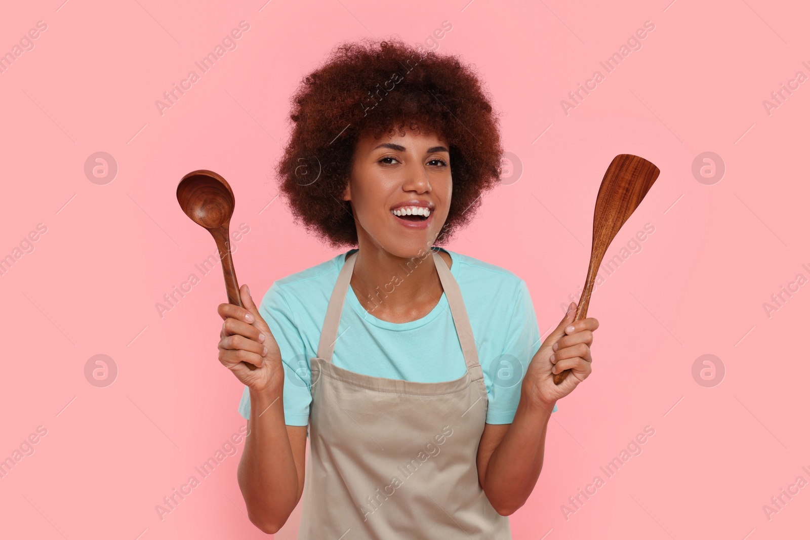 Photo of Happy young woman in apron holding spoon and spatula on pink background