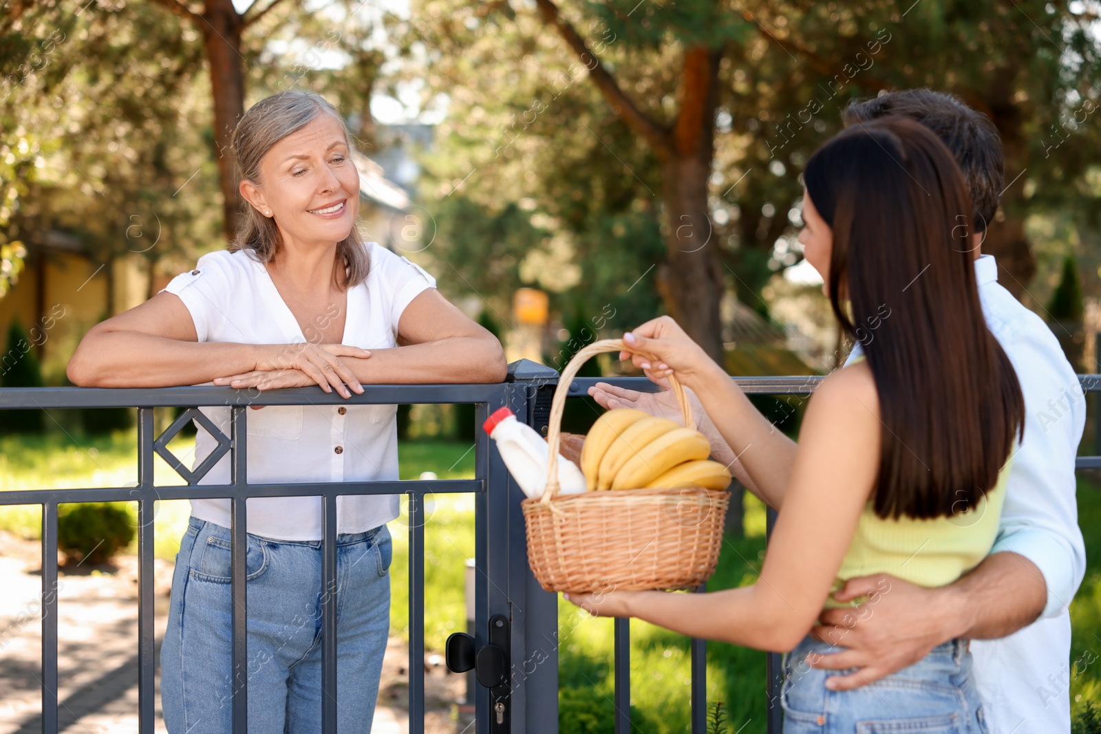 Photo of Friendly relationship with neighbours. Young couple with wicker basket of products treating senior woman near fence outdoors