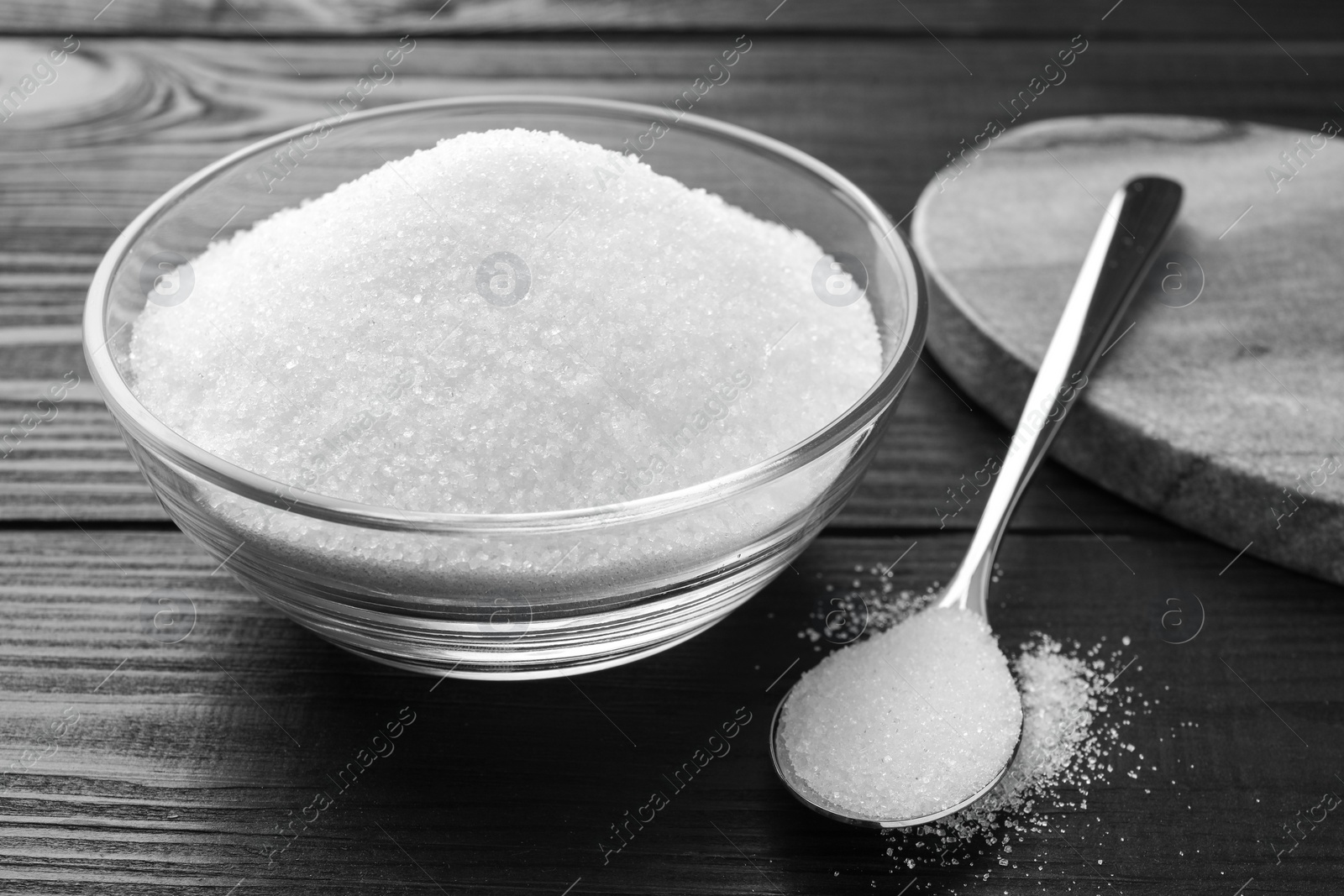 Photo of Granulated sugar in bowl and spoon on black wooden table, closeup