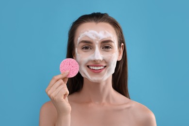 Happy young woman washing her face with sponge on light blue background