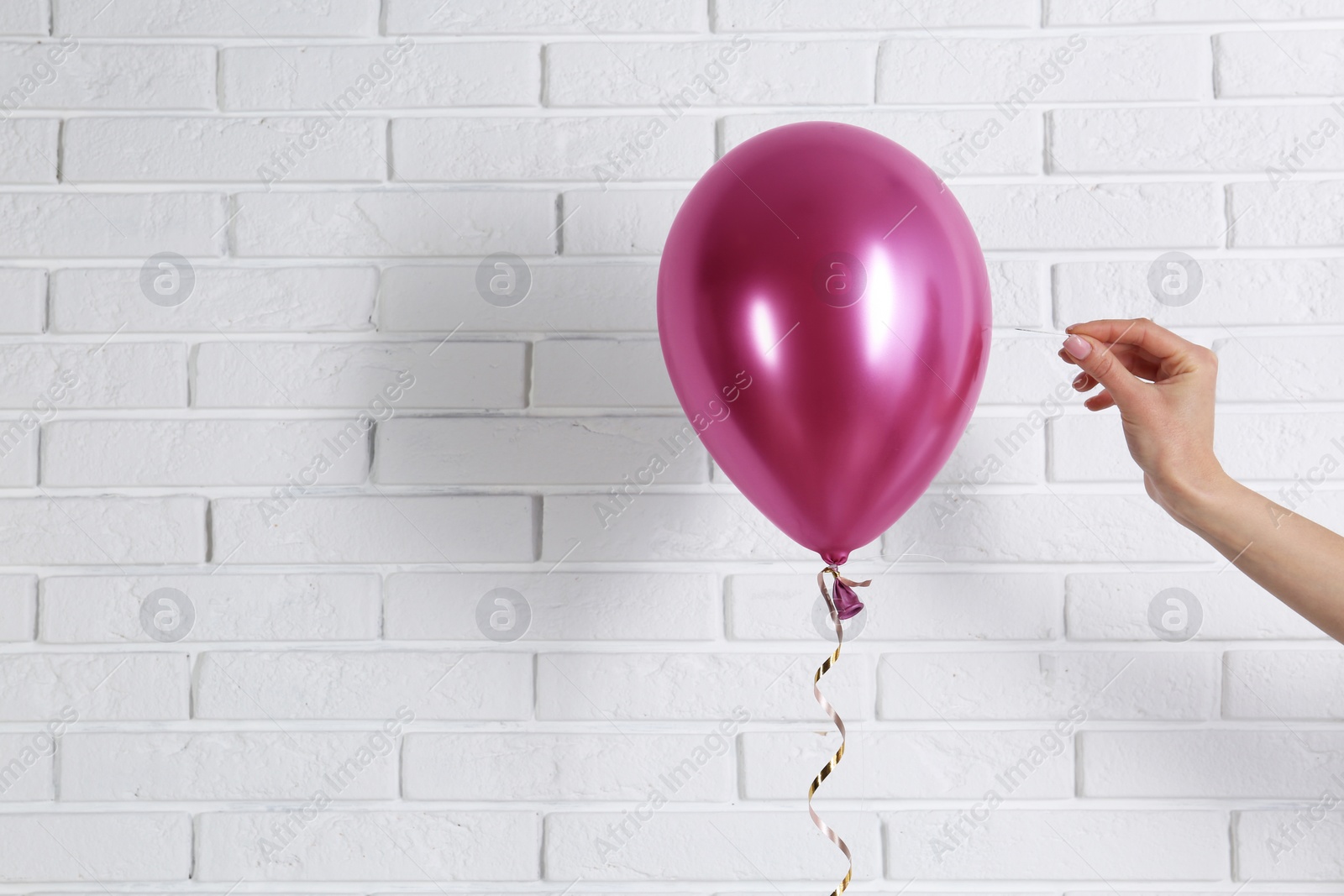Photo of Young woman piercing bright balloon near brick wall, space for text
