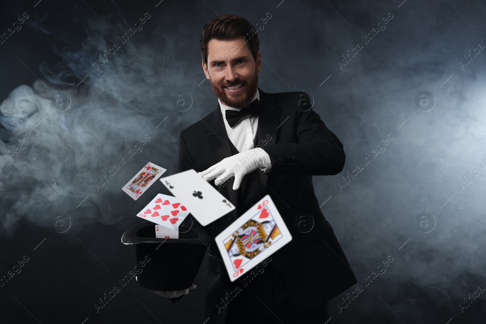 Image of Smiling magician showing trick with cards and top hat on dark background