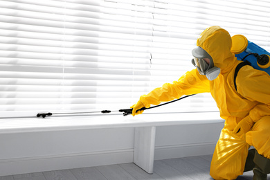 Photo of Male worker in protective suit spraying insecticide on window sill indoors. Pest control