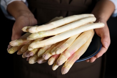 Photo of Woman holding plate with fresh white asparagus on black background, closeup