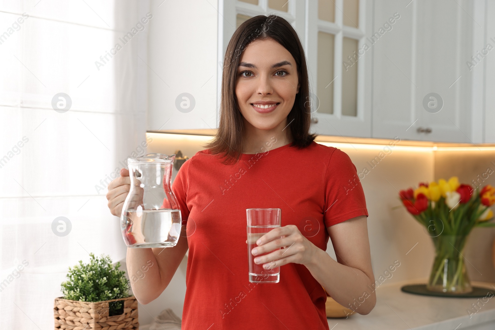 Photo of Young woman with jug and glass of water in kitchen
