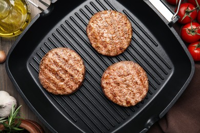 Photo of Grill pan with tasty fried hamburger patties and seasonings on wooden table, flat lay