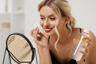 Smiling woman removing makeup with cotton pad in front of mirror indoors