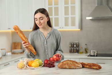 Photo of Woman with baguette, apple and string bag of fresh fruits at light marble table in kitchen