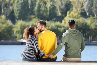 Photo of Woman holding hands with another man behind her boyfriend's back on pier near river. Love triangle