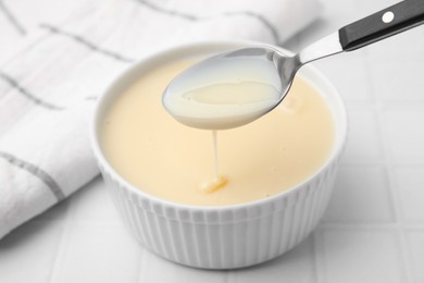 Photo of Condensed milk flowing down from spoon into bowl on white tiled table, closeup