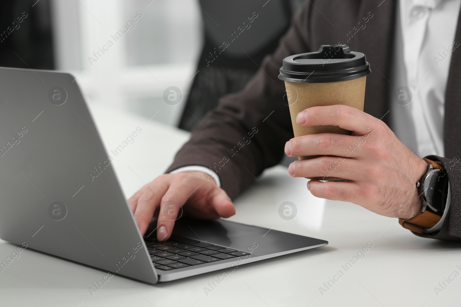 Photo of Man with cup of coffee working on laptop at white desk in office, closeup