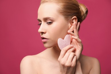 Beautiful young woman doing facial massage with gua sha tool on pink background, closeup