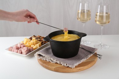 Dipping piece of ham into fondue pot with tasty melted cheese at white table, closeup