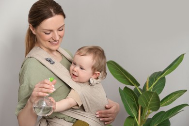 Photo of Mother spraying houseplant with water while holding her child in sling (baby carrier) at home