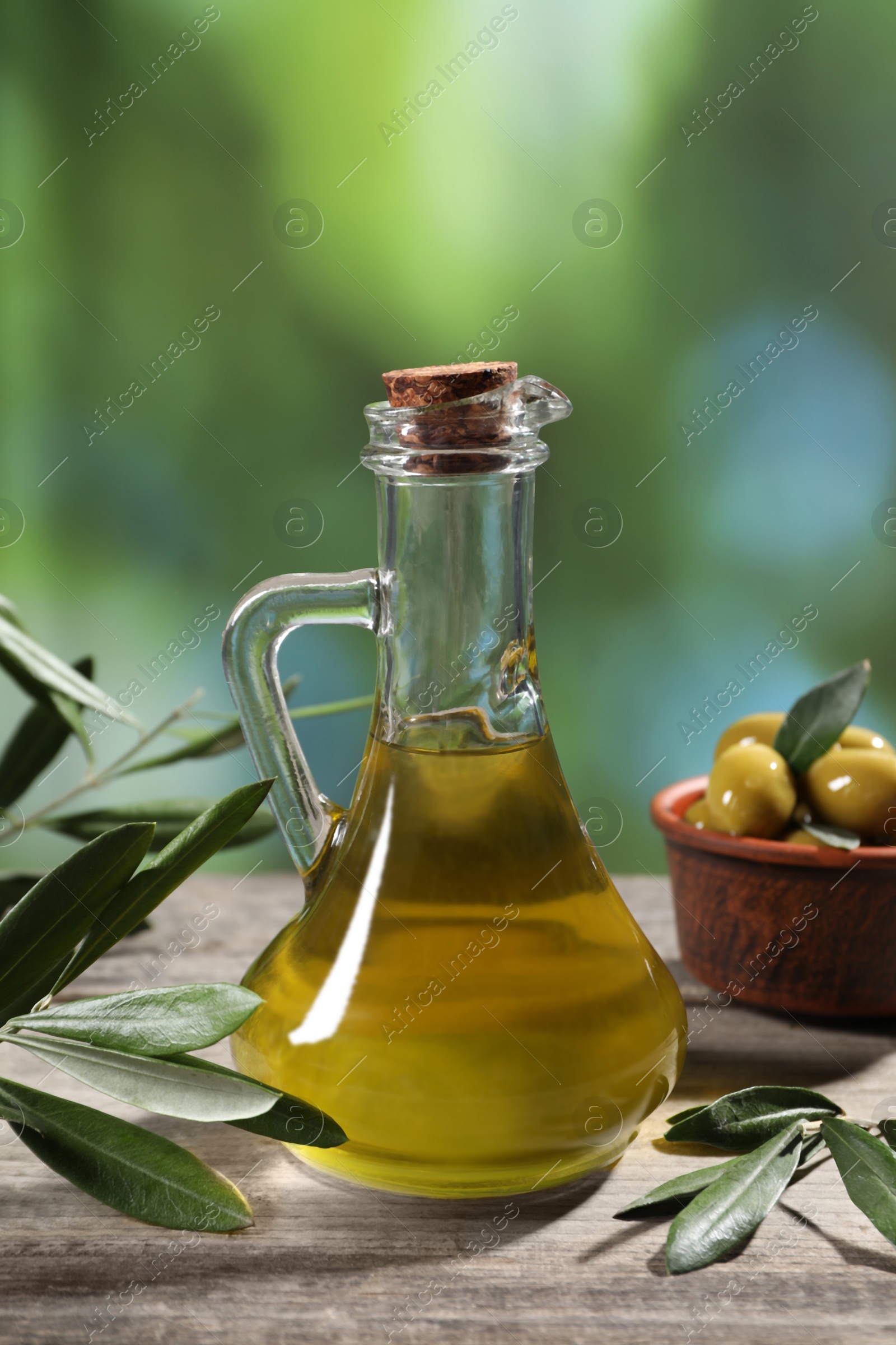 Photo of Jug of cooking oil, olives and green leaves on wooden table against blurred background