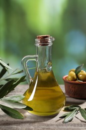 Jug of cooking oil, olives and green leaves on wooden table against blurred background