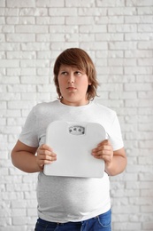 Emotional overweight boy with floor scales near white brick wall