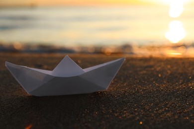 Photo of White paper boat on sand near sea at sunset, closeup. Space for text