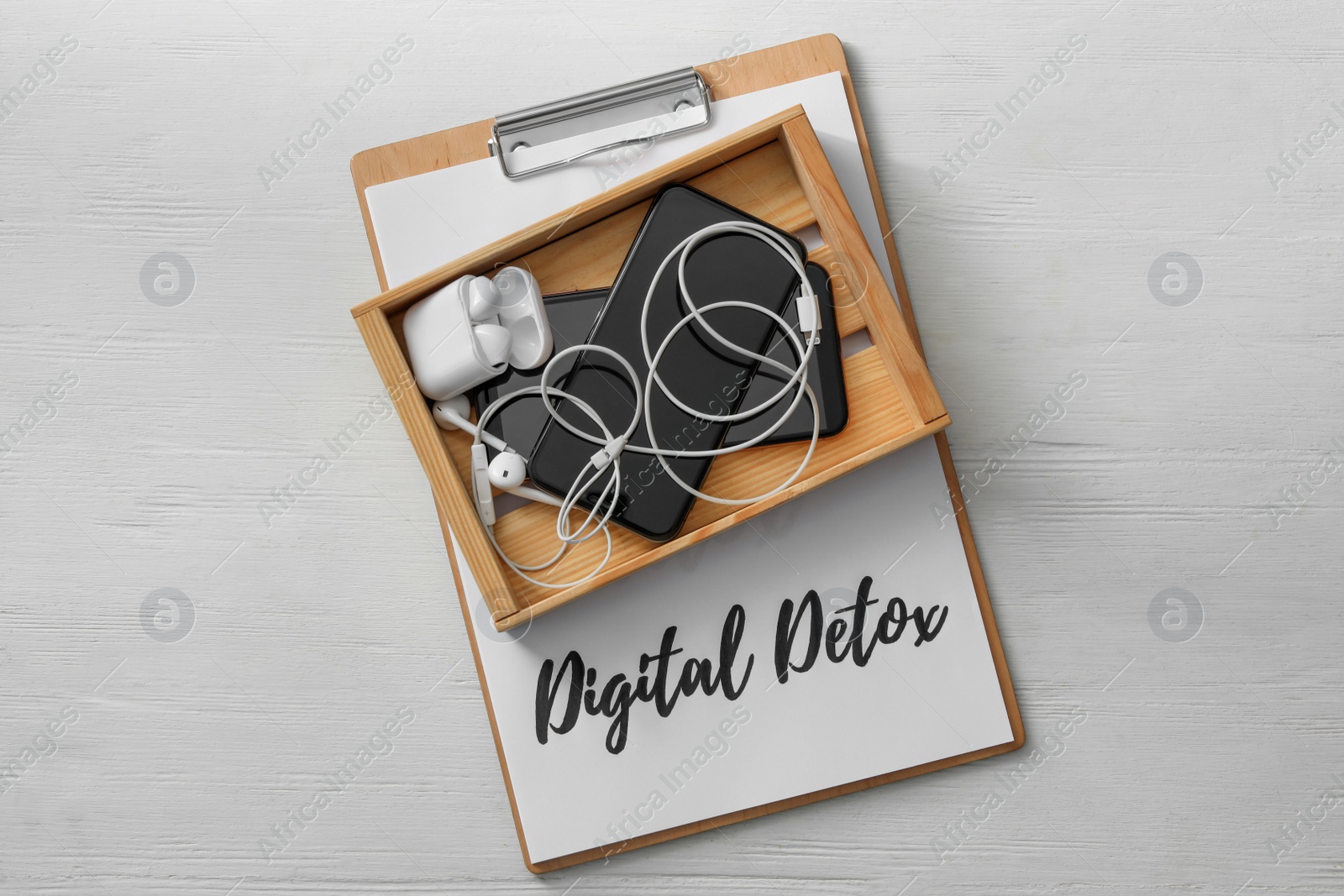 Photo of Different gadgets in crate and clipboard with words DIGITAL DETOX on white wooden background, top view