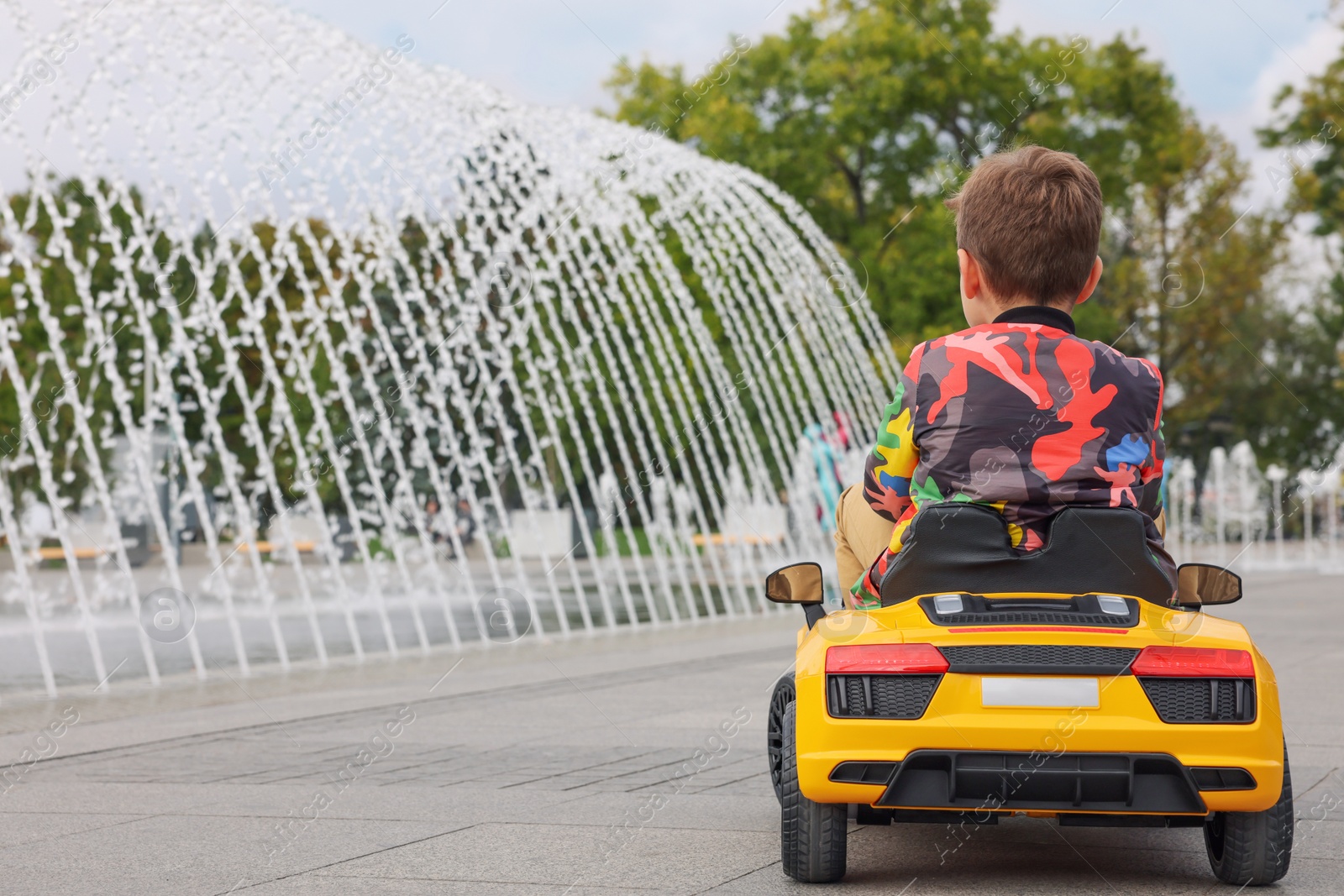 Photo of Cute little boy driving children's car near fountain on city street, back view. Space for text