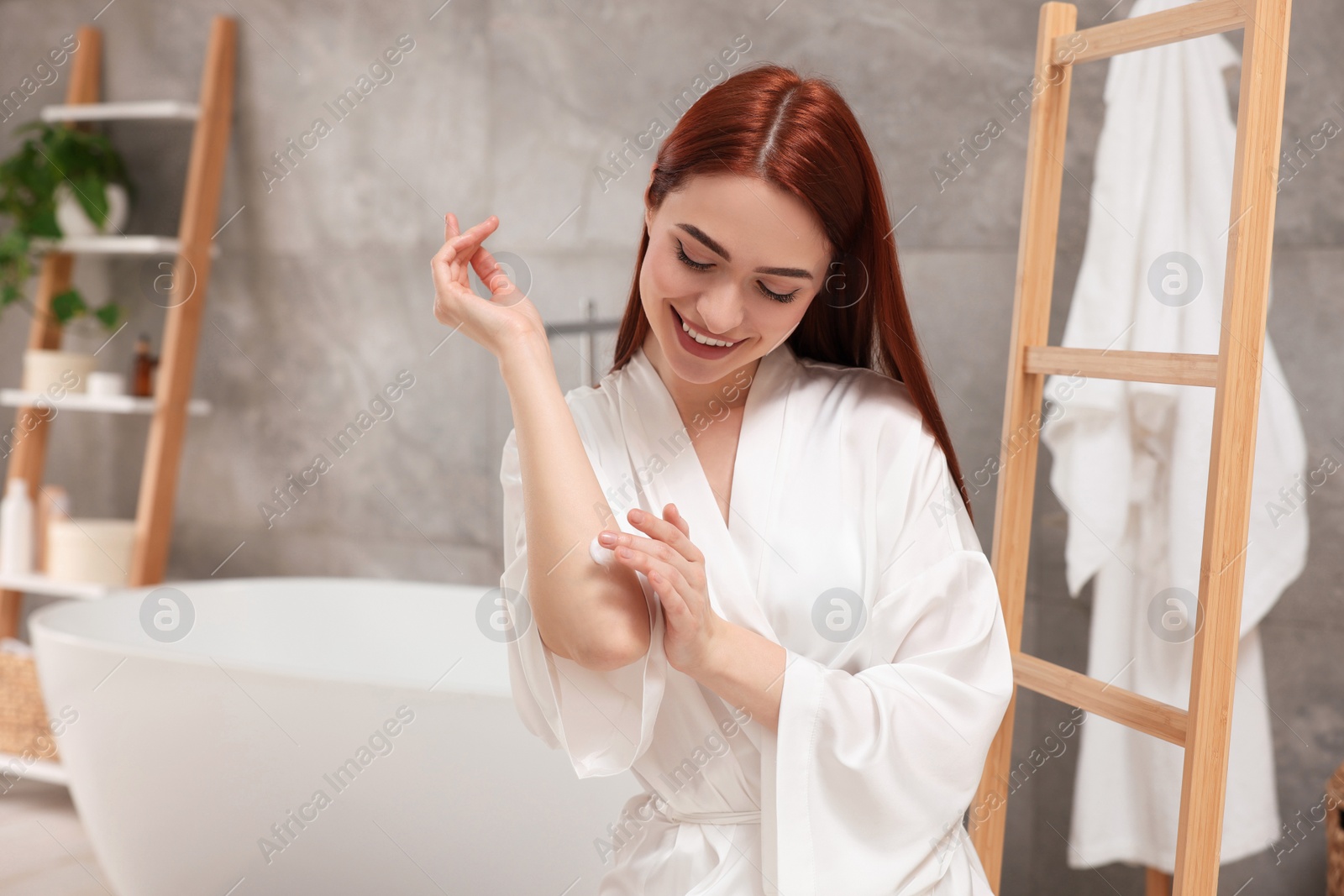 Photo of Beautiful young woman applying body cream onto elbow in bathroom, space for text