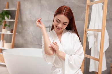 Beautiful young woman applying body cream onto elbow in bathroom, space for text