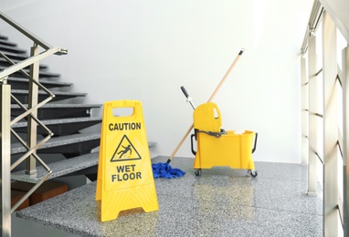 Photo of Safety sign with phrase "CAUTION WET FLOOR" and mop bucket on stairs. Cleaning tools